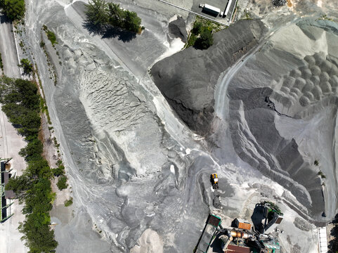 Aerial view of an industrial quarry with large piles of grey gravel, heavy machinery, and surrounding green trees Caprie, Piedmont, Italy.