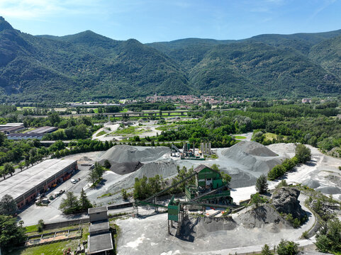 Aerial view of an industrial quarry with gravel piles and machinery nestled in a green valley against the backdrop of the Alps in Caprie, Piedmont, Italy.