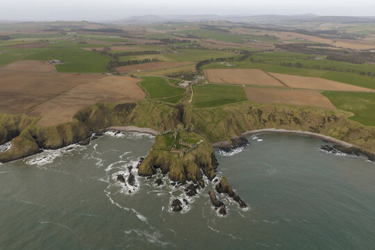 Aerial view of Dunnottar Castle, a ruined medieval fortress on a rocky cliff edge overlooking the North Sea with surrounding green fields in Stonehaven, Scotland, United Kingdom.