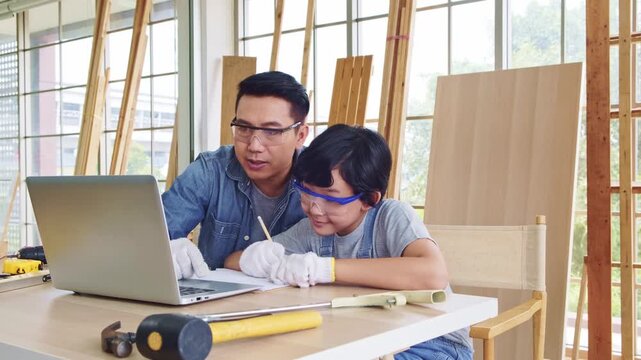 An adult and child wearing safety glasses learn woodworking with online guidance, using a laptop at a workshop table surrounded by tools and wooden boards.