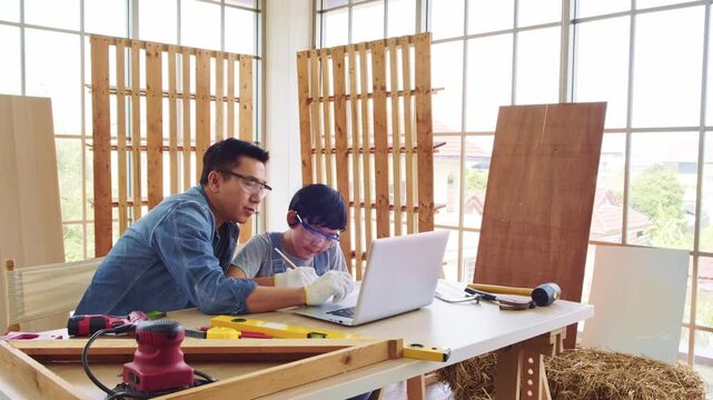Cheerful Asian carpentry father discusses and teaches his son about construction instruments on laptop at the home workshop studio. Construction work and repairs. Carpentry and family concept.