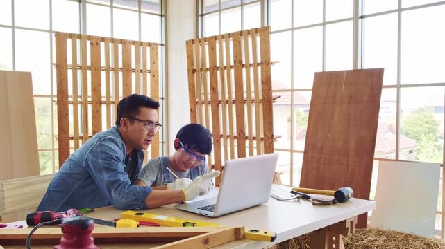 Cheerful Asian carpentry father discusses and teaches his son about construction instruments on laptop at the home workshop studio. Construction work and repairs. Carpentry and family concept.