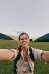 Young woman outdoors takes a selfie while hiking through a lush green valley, smiling with wind in...