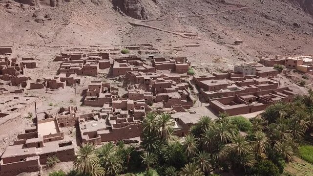 Aerial drone footage of a traditional desert village surrounded by a palm oasis and arid mountains in Morocco. Strong contrast between human settlement and natural environment, ideal for documentary,