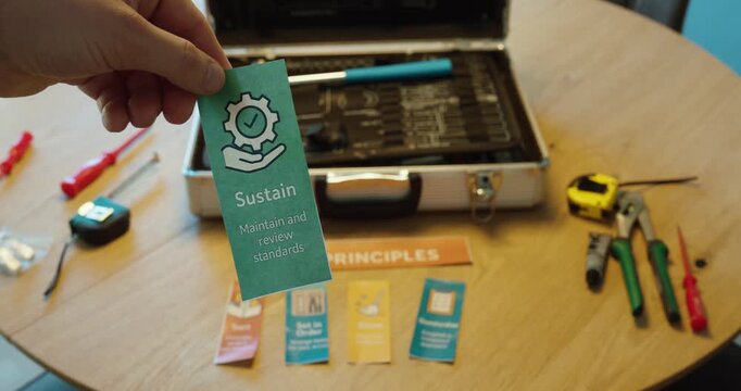 Hand holding a &ldquo;Sustain&rdquo; 5S principle card with tools and workspace in background, representing standardization and continuous improvement.