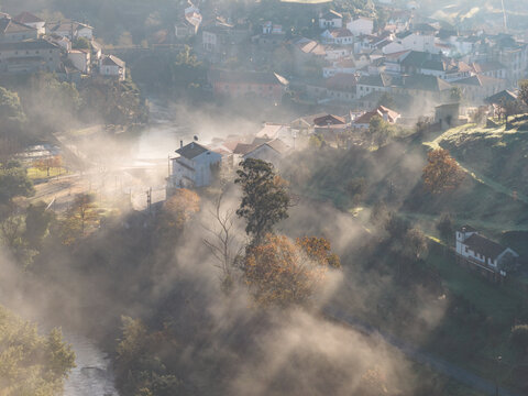Aerial view of the historic village of Avo shrouded in morning mist with sunlight rays piercing through the fog and lush green hills Oliveira do Hospital, Coimbra District, Portugal.