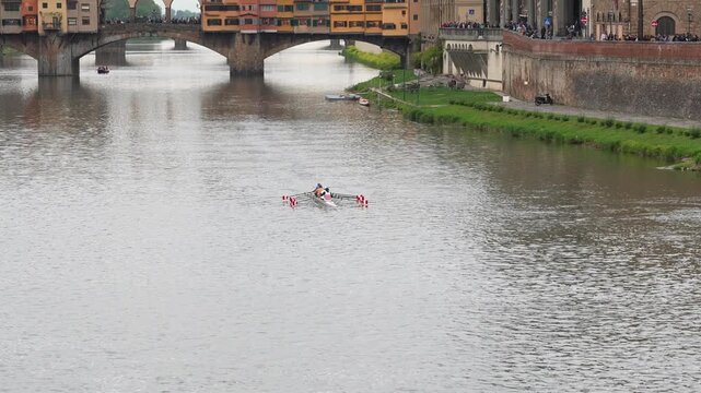 Wide shot of rowers gliding on the Arno River beneath the iconic Ponte Vecchio in Florence, Italy. Overcast daylight enhances historic architecture, vibrant water activity, and scenic city life.