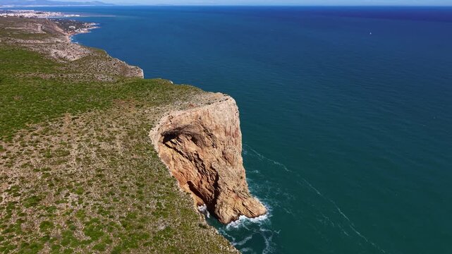 Dramatic sea cliff with natural cave arch rises from turquoise Mediterranean waters along a rugged coastline with green scrubland stretching toward a distant coastal town