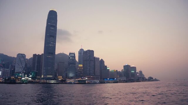 Handheld wide shot from a moving boat of Hong Kong island skyline at dusk, featuring IFC tower, Central waterfront, and Victoria Peak, with calm harbor water in foreground, cinematic and atmospheric