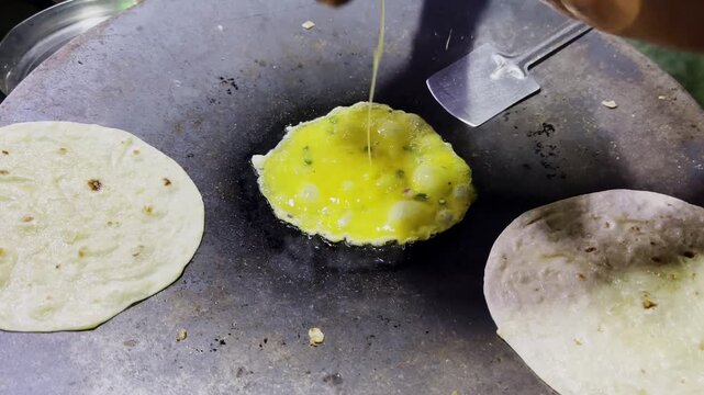 Egg mixture being poured and cooked on hot pan before chapati is placed over omelet, traditional street food preparation with rustic kitchen atmosphere!