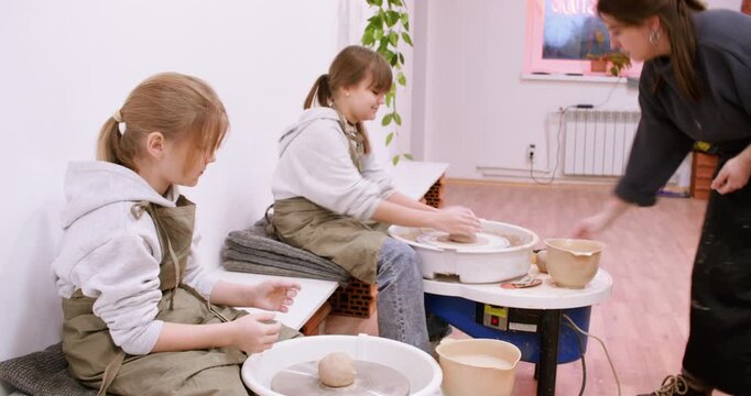 Children pottery. Artisan master class. Professional woman teaching kids girls handmade ceramic using electric wheel in workplace.