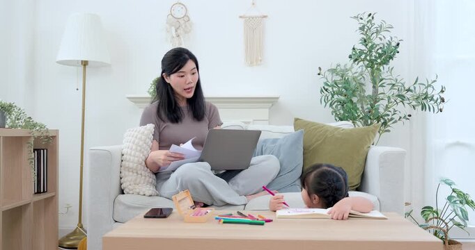 Asian single mother sitting on sofa with laptop and paper, watching preschool daughter draw in workbook at table, modern home education routine, childcare support, balanced family lifestyle, care