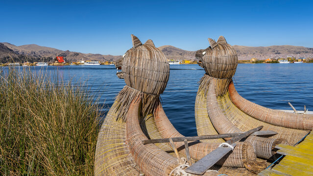 Traditional balsa de totora boat off the coast of the Uros Floating Island. There are decorations in the form of puma heads on the bow of the boat. The oars are on deck. Ripples on the blue water. 
 