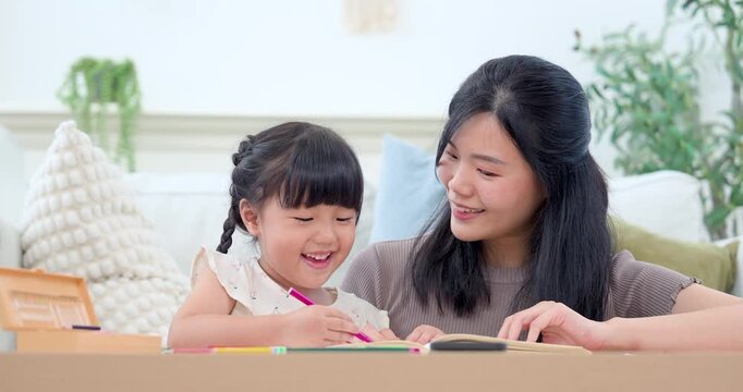 Asian single mother shares drawing time with preschool daughter at home table, encouraging creativity, bonding, and early childhood learning as child smiles while practicing with colorful pencil.