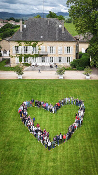 La Chapelle-de-Guinchay, France - 01 June 2024: Aerial view of a large group of people forming a heart shape on a green lawn in front of a historic manor house.