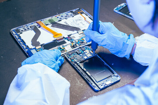 young female electronics technician carefully repairs a smartphone at her workstation. She uses professional precision screwdrivers and tweezers under a bright workspace light. 