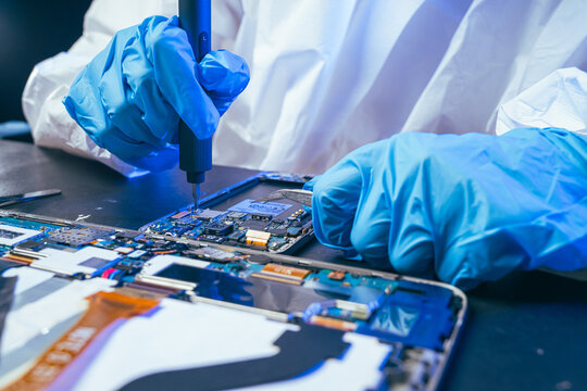 young female electronics technician carefully repairs a smartphone at her workstation. She uses professional precision screwdrivers and tweezers under a bright workspace light. 