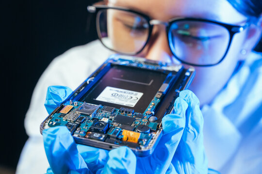 young female electronics technician carefully repairs a smartphone at her workstation. She uses professional precision screwdrivers and tweezers under a bright workspace light. 