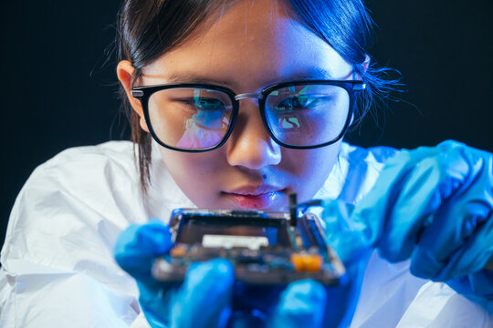 young female electronics technician carefully repairs a smartphone at her workstation. She uses professional precision screwdrivers and tweezers under a bright workspace light. 