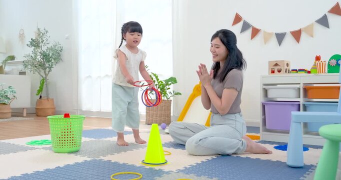 Asian single mother claps for preschool daughter after ring toss attempt in home playroom, celebrating success and building confidence, coordination, and happy bonding through educational floor game.
