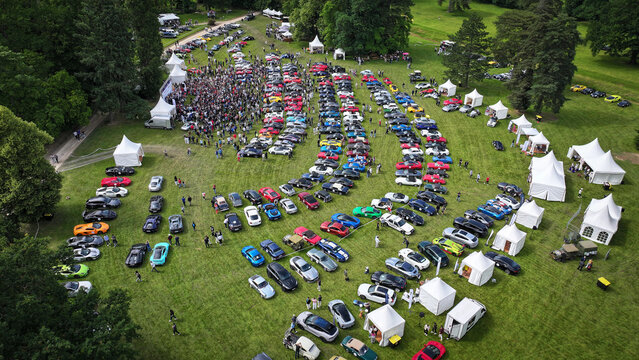 La Chapelle-de-Guinchay, France - 01 June 2024: Aerial view of a car show with rows of colorful sports cars on a green lawn with people walking among white tents and lush trees.