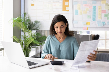 Fototapeta na wymiar Focused professional woman reviewing financial documents, holds invoice, preparing report or budget summary, using calculator for expenses and incomes calculations seated at workplace desk with laptop