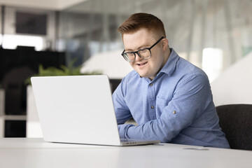 Fototapeta na wymiar Smiling professional male office manager with Down syndrome working on laptop, review tasks to-do, engaged in administrative remote workflow seated at desk in contemporary workspace. Inclusivity, tech