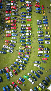 Aerial view of colorful sports cars and vintage vehicles parked in rows on a green field during a car show with people walking around La Chapelle-de-Guinchay, Bourgogne-Franche-Comte, France.