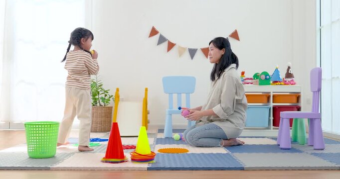 Asian single mother guides preschool daughter holding yellow ball near basket and cone, building throwing skill, hand eye coordination, confidence, and physical development through active home play.