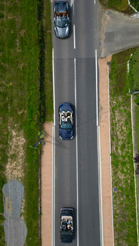 Aerial view of three convertible cars with people inside driving along a paved road bordered by green grass and vegetation in La Chapelle-de-Guinchay, Bourgogne-Franche-Comte, France.