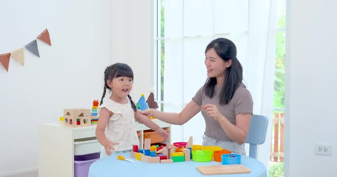 Asian single mother cheering preschool daughter during colorful block play at round table, energetic home development activity encouraging confidence, communication, motor skill, joyful bonding