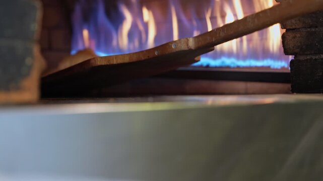 Cinematic close-up of fresh pita bread baking in a traditional stone oven with blue flames. A wooden peel enters the frame, perfect for restaurant marketing and culinary storytelling.