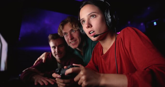 Closeup young adult female gamer wearing headset, holding game controller, playing videogame in cybercafe with male friends, focused on gameplay, interacting during multiplayer session in gaming club