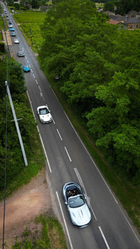 Aerial view of a convoy of luxury sports cars with people driving along a narrow road through lush green trees and vineyards in La Chapelle-de-Guinchay, Bourgogne-Franche-Comte, France.