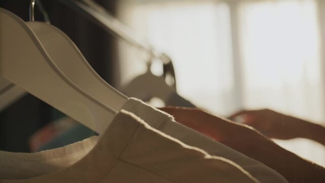 Person sifts through clothes on hangers near window with soft light.