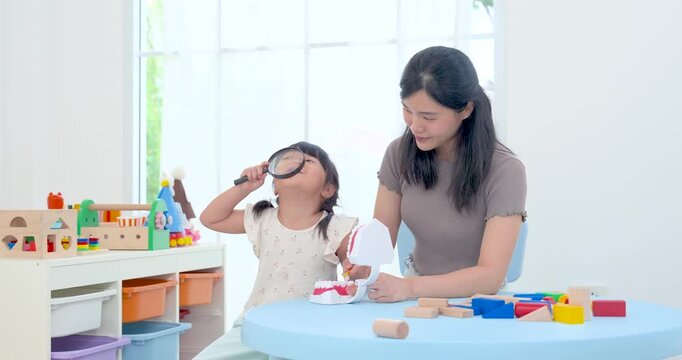 Asian single mother guiding preschool daughter using magnifying glass with dental model at round table, creative home learning play encouraging observation, tooth care awareness, bonding, development
