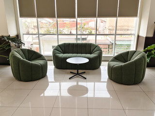 Fototapeta premium Modern green sofa set sits beneath large windows. Circular table rests at center, inviting conversation. Bright tiles reflect light, enhancing open space. Potted plants add natural touch