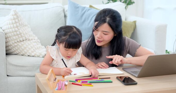 Young Asian single mother points toward workbook while little daughter sketches with orange pencil near laptop and crayon on table, encouraging art practice, concentration, patience, and learning.