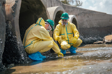 Scientists in hazardous material protection suits conduct wastewater testing near concrete drainage tunnels. Industrial pollution monitoring and improve water quality management. © Wanwajee