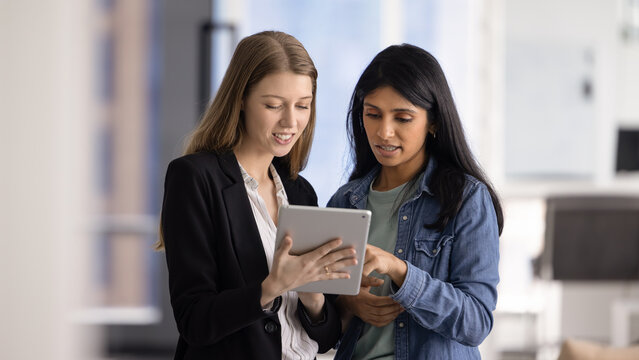 Two young attractive multiethnic women workmates, team members using innovative tools on digital tablet in modern office, discussing data or project details. Collaboration, professional teamwork, tech