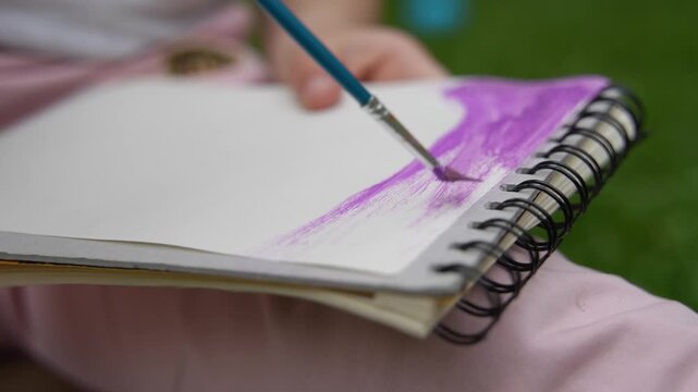 Close up of child painting with watercolor brush in sketchbook outdoors