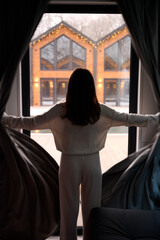 Obraz premium Woman opening curtains and looking through window at cozy illuminated cabins, representing winter travel, hospitality, home comfort, festive season, and peaceful lifestyle moments