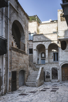 View of historic stone buildings with arched entrances, external staircases, and balconies under a clear blue sky in the old town of Polignano a Mare, Apulia, Italy.