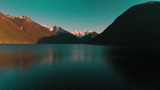 Expansive view across dark calm rippling lake water facing majestic rugged mountain range featuring snow capped peaks glowing intensely with bright golden sunset light ascending approach aerial drone
