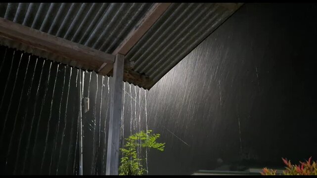 Heavy rain falling from a corrugated roof edge at night, backlit by a bright light source creating a beautiful lens flare and glowing water droplets.