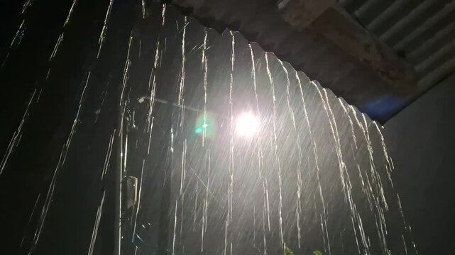Heavy rain falling from a corrugated roof edge at night, backlit by a bright light source creating a beautiful lens flare and glowing water droplets.