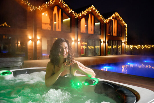 Smiling woman relaxing in jacuzzi beside illuminated modern lodge, representing leisure, self care, indulgence and aspirational vacation living