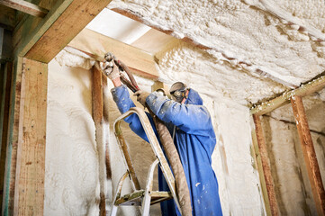 Builder applying spray foam insulation along wall and ceiling junction inside wooden frame home....
