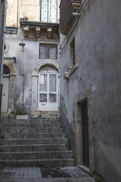 View of a narrow alleyway with stone steps and weathered historic building facades in Noto, Sicily, Italy.
