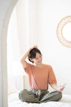 ベッドにあぐらをかいて座りながらスマホを見ているショートカットの女性